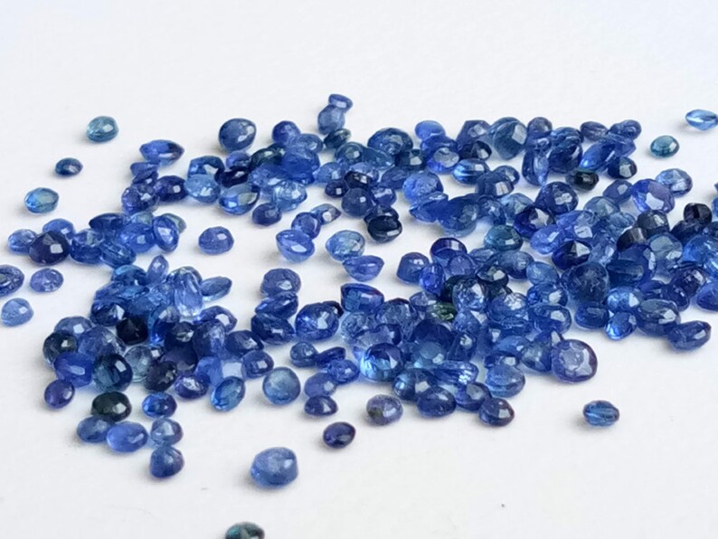 May include: A loose pile of small, faceted, blue gemstones. The gemstones are scattered on a white surface.