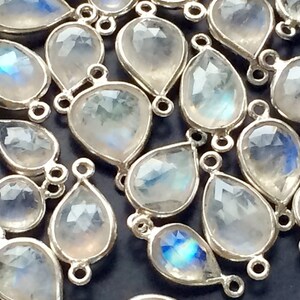 Puede incluir: Primer plano de un grupo de colgantes de piedra preciosa en forma de lágrima de color blanco nacarado con anillas de salto plateadas. Las piedras preciosas tienen un brillo iridiscente sutil.