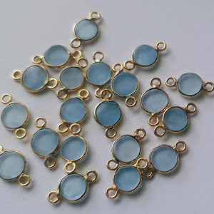 Pode incluir: Uma coleção de 18 contas de pedra preciosa azul claro com laços de conexão de metal dourado. As contas são redondas e têm um acabamento liso e polido.