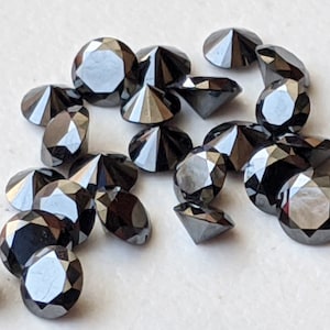 May include: A collection of black round faceted gemstones, some with a pointed top, scattered on a white surface.
