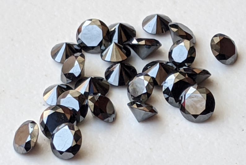 May include: A collection of black round faceted gemstones, some with a pointed top, scattered on a white surface.
