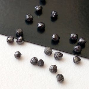 May include: A collection of black rough diamonds, also known as carbonado diamonds, scattered on a black and white surface. The diamonds are various sizes and shapes.