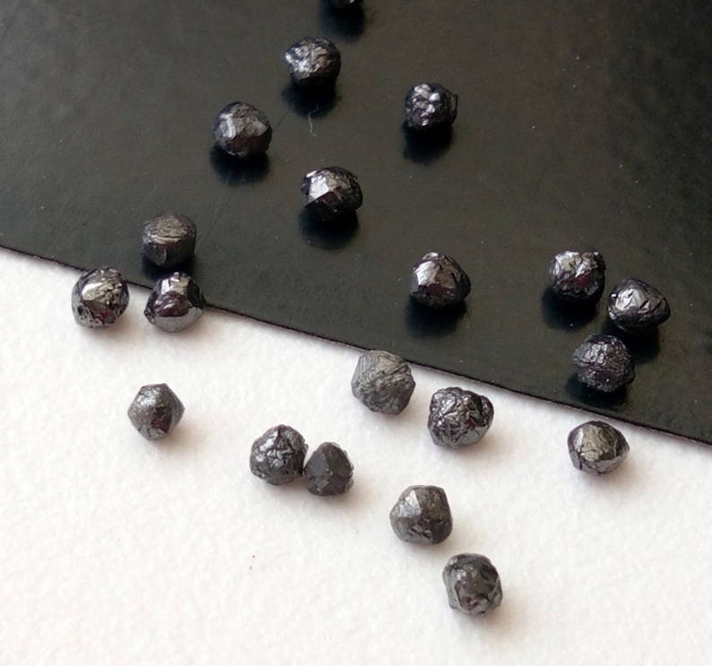 May include: A collection of black rough diamonds, also known as carbonado diamonds, scattered on a black and white surface. The diamonds are various sizes and shapes.