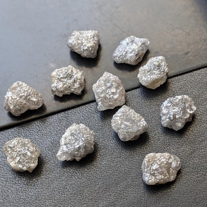 K&ouml;nnte beinhalten: Eine Sammlung von zw&ouml;lf rohen, unpolierten grauen Diamanten. Die Diamanten sind in verschiedenen Gr&ouml;&szlig;en und Formen und alle auf einer grauen Oberfl&auml;che zusammen.