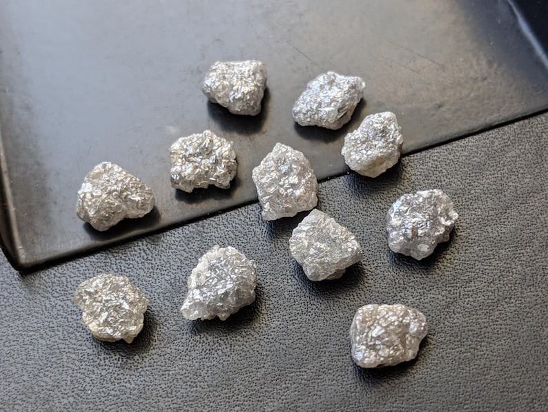 K&ouml;nnte beinhalten: Eine Sammlung von zw&ouml;lf rohen, unpolierten grauen Diamanten. Die Diamanten sind in verschiedenen Gr&ouml;&szlig;en und Formen und alle auf einer grauen Oberfl&auml;che zusammen.