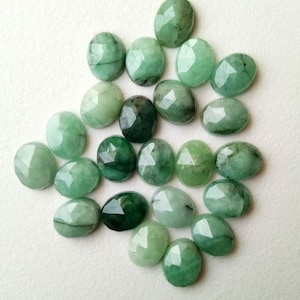 May include: A collection of 20 faceted emerald green gemstones, each with a unique shape and size. The stones are arranged in a loose pile on a white background.