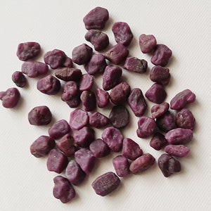 May include: A collection of small, rough, purple ruby gemstones. The stones are irregularly shaped and have a natural, unpolished finish.