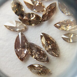 May include: A collection of brown marquise-shaped diamonds, also known as navette diamonds, are displayed on a white surface. The diamonds are faceted and have a slight golden hue.