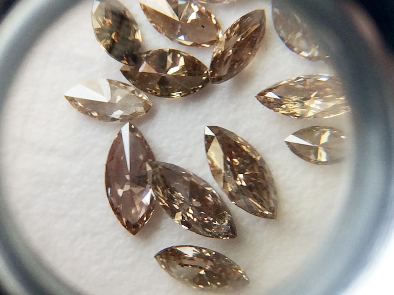 May include: A collection of brown marquise-shaped diamonds, also known as navette diamonds, are displayed on a white surface. The diamonds are faceted and have a slight golden hue.