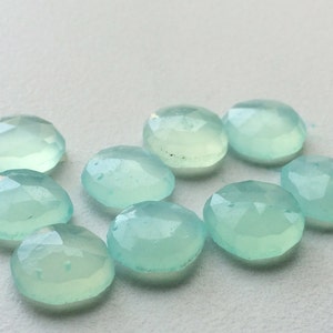 May include: A collection of nine light blue, oval-shaped gemstones. Each gem has a faceted surface, catching the light and creating a sparkling effect. The gems are arranged on a white surface, showcasing their color and clarity. Ideal for jewelry making.