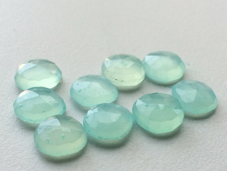 May include: A collection of nine light blue, oval-shaped gemstones. Each gem has a faceted surface, catching the light and creating a sparkling effect. The gems are arranged on a white surface, showcasing their color and clarity. Ideal for jewelry making.