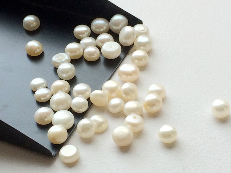 May include: A collection of white round pearls of various sizes, scattered on a white surface and spilling from a black tray.