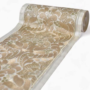 Puede incluir: Un rollo de tela decorativa con un diseño floral dorado sobre un fondo crema. La tela está enrollada, revelando el intrincado diseño y la textura. Los bordes están acabados con un borde color crema.