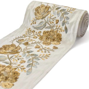 Könnte beinhalten: Eine Rolle cremefarbenen Stoffes mit gold- und silberfarbener Blumenstickerei. Das Design zeigt große, detaillierte Blumen und zarte Blätter. Der Stoff ist gerollt und zeigt die sich wiederholende Natur des Musters, geeignet für Bastel- und Nähprojekte.
