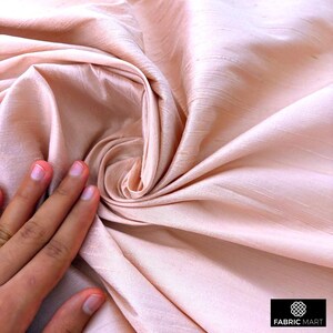 May include: A close-up of a soft, peach-colored fabric with a silky texture. The fabric is draped and folded, creating a smooth, flowing look.