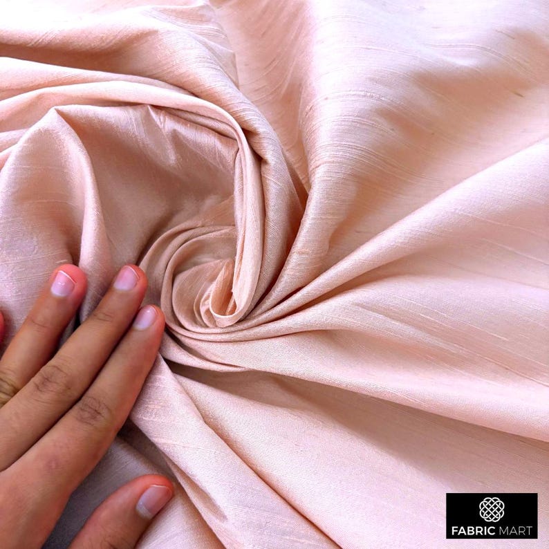 May include: A close-up of a soft, peach-colored fabric with a silky texture. The fabric is draped and folded, creating a smooth, flowing look.