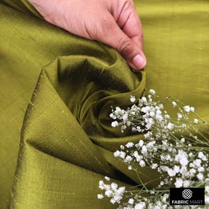 Puede incluir: Primer plano de una tela de seda verde con un brillo sutil. La tela est&aacute; drapeada y doblada, creando una textura suave y fluida. Una ramita de flores blancas a&ntilde;ade un toque delicado a la imagen.