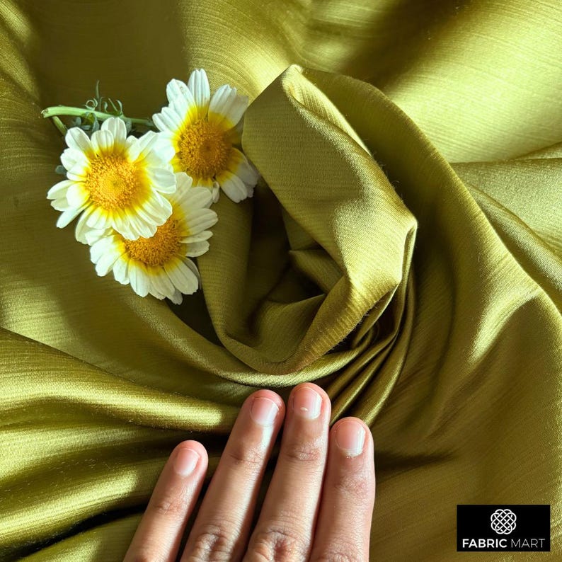 May include: A close-up of a hand resting on a smooth, olive green fabric. The fabric is folded and draped, creating a soft, flowing texture. Three white daisies with yellow centers are placed on the fabric.
