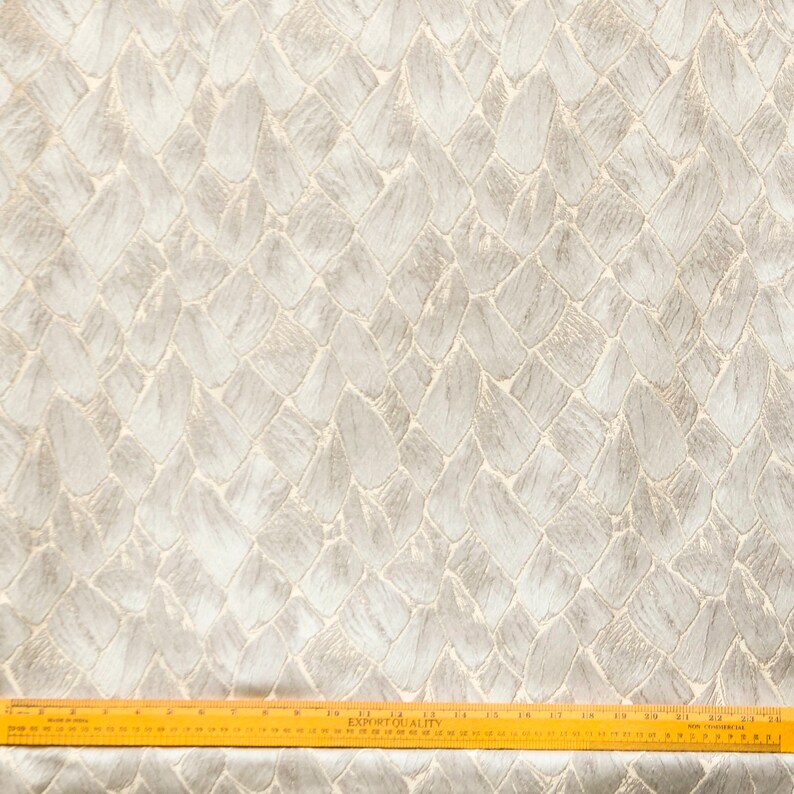 May include: Close-up of a white fabric with a textured pattern of overlapping gray shapes. The fabric has a subtle sheen and a woven texture. A wooden ruler is placed at the bottom of the image, with the text "EXPORT QUALITY" visible.