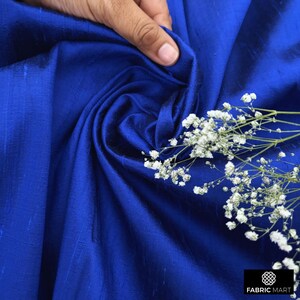 May include: A close-up of a royal blue silk fabric with a subtle sheen. The fabric is draped and folded, creating soft folds and wrinkles. A sprig of white baby's breath flowers is placed on the fabric.