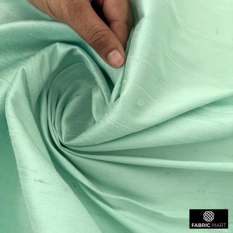 May include: A light green silk fabric with a smooth, silky texture. The fabric is folded and draped, showcasing its drape and flow.