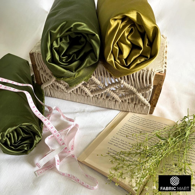 May include: Two rolls of fabric, one green and one gold, are displayed on a woven macrame shelf. A measuring tape and a book are on a white surface below the shelf.