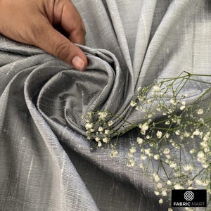 May include: Close-up of a silver silk fabric with a subtle sheen. The fabric is draped and folded, creating a soft, flowing texture. A sprig of white flowers adds a delicate touch.