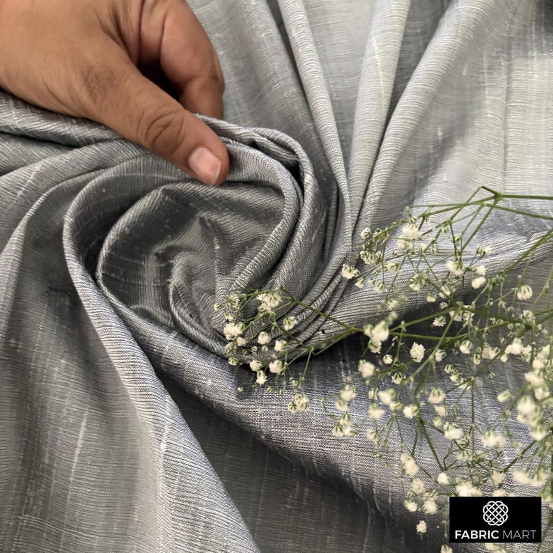 May include: Close-up of a silver silk fabric with a subtle sheen. The fabric is draped and folded, creating a soft, flowing texture. A sprig of white flowers adds a delicate touch.