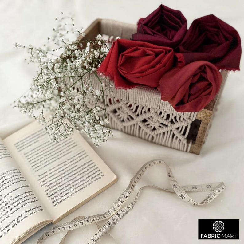 May include: A wooden box with a white macrame design holds three red and burgundy fabric squares. A measuring tape and a book are on a white surface. The text "FABRIC MART" is visible in the lower right corner of the image.