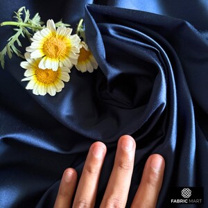 May include: A close-up of a deep blue satin fabric with a hand resting on the surface. The fabric is draped and folded, creating a soft, luxurious look. White daisies are placed on the fabric, adding a touch of elegance.
