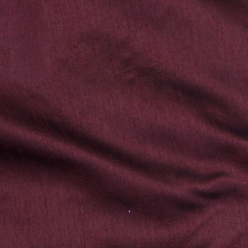 May include: A close-up of a deep burgundy fabric with a smooth, slightly textured surface. The fabric is folded and draped, creating soft folds and wrinkles.