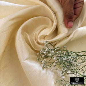 May include: A close-up of a smooth, ivory-colored fabric with a subtle sheen. The fabric is draped and folded, creating soft curves and folds. A sprig of white flowers adds a delicate touch to the image.