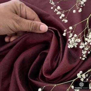 May include: Close-up of a deep burgundy silk fabric with a subtle sheen. The fabric is draped and folded, creating soft folds and curves. A sprig of white flowers is placed on top of the fabric.