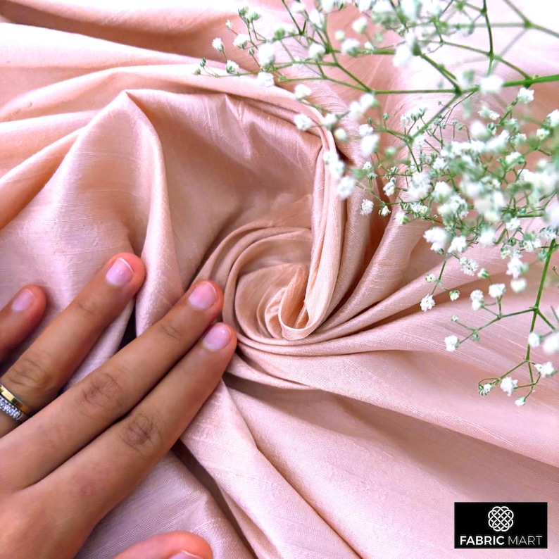May include: A close-up of a soft, peach-colored fabric with a delicate, silky texture. The fabric is draped and folded, creating a smooth, flowing effect. The fabric is accented with a sprig of white baby's breath flowers.