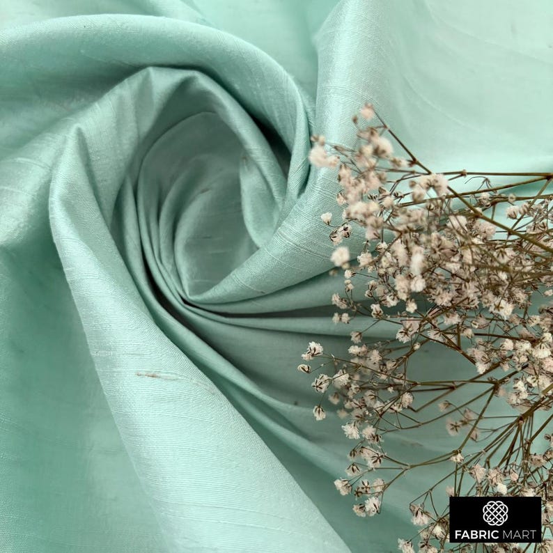 May include: A light blue silk fabric with a subtle sheen. The fabric is draped and folded, creating soft folds and curves. A sprig of white flowers is placed on the fabric.