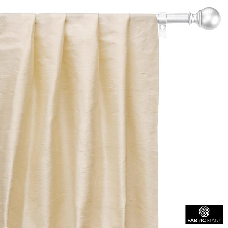 May include: A close-up of a cream-colored curtain with pleats hanging from a white curtain rod with a silver finial.