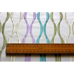 Puede incluir: Tela blanca con un patr&oacute;n repetitivo de l&iacute;neas onduladas en tonos de verde, morado y azul. Una regla de madera se coloca encima de la tela, con los n&uacute;meros del 1 al 20 visibles.