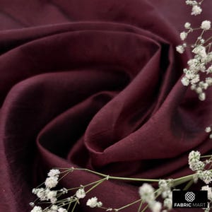 May include: Close-up of a deep burgundy fabric with a subtle sheen. The fabric is draped and folded, creating soft folds and wrinkles. Small white flowers are scattered across the fabric.