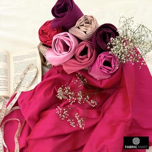 May include: A collection of pink, red, and purple fabric swatches rolled up and arranged in a basket. The fabric is displayed on a pink background with a book and measuring tape.