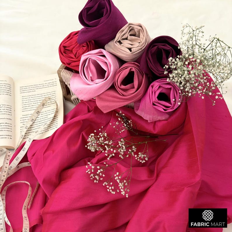 May include: A collection of pink, red, and purple fabric swatches rolled up and arranged in a basket. The fabric is displayed on a pink background with a book and measuring tape.