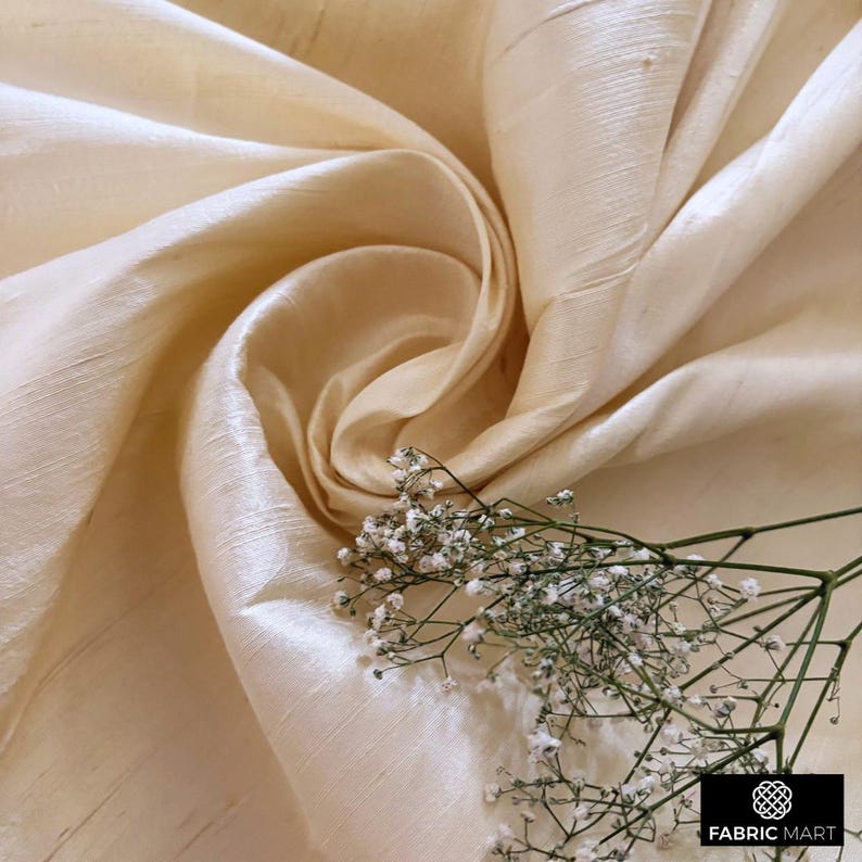 May include: Cream colored silk fabric with a subtle sheen. The fabric is draped and folded to show its texture. A sprig of white flowers is placed on the fabric.