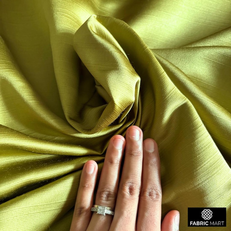 May include: Close-up of a smooth, olive green fabric with a subtle sheen. The fabric is draped and folded, creating soft curves and folds. A hand with a ring is resting on the fabric.
