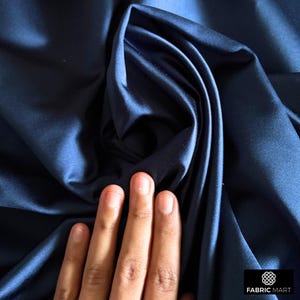 May include: Close-up of a deep blue satin fabric, showcasing its smooth texture and drape. The fabric is folded and held by a hand, highlighting its luxurious feel.