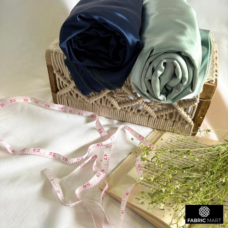 May include: Two rolls of fabric, one dark blue and one light blue, are displayed in a woven basket. A pink measuring tape is lying on a white surface next to a book and a bouquet of white flowers.