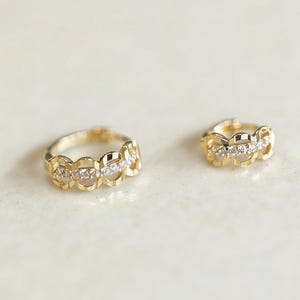 Può includere: Un paio di orecchini a cerchio color oro. Ogni orecchino presenta una serie di forme traforate collegate, con piccole pietre trasparenti incastonate al centro. Gli orecchini sono esposti su una superficie chiara.