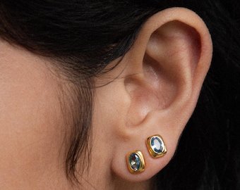 May include: Two gold stud earrings featuring rectangular settings with rounded corners. Each earring contains a central oval-shaped blue gemstone. The earrings are positioned on an earlobe, with one placed slightly above and behind the other.