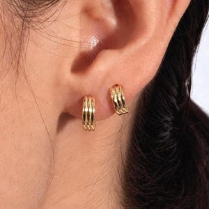 May include: Two small gold hoop earrings worn in the earlobe. Each hoop features a triple-band design with a polished, reflective finish. The earrings are circular in shape and sit closely against the skin of the ear, showing a metallic texture.