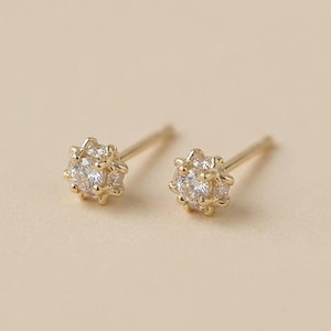 May include: A pair of gold-colored stud earrings. Each earring features a central, clear gemstone surrounded by smaller, clear gemstones, creating a starburst design. The earrings are set against a neutral background.