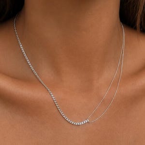 May include: A silver-toned necklace featuring a dual-strand design. One side consists of a thicker, textured curb chain, while the other side transitions into a single, thinner, smooth cable chain, creating an asymmetrical layered appearance.