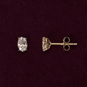 May include: A pair of stud earrings featuring oval-cut clear stones set in yellow gold. One earring is shown from the front, displaying the faceted stone held by four prongs, while the other is shown from the side, revealing the gold post and backing.